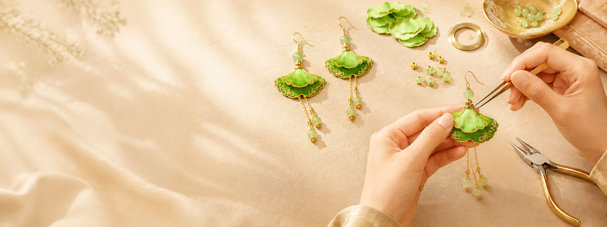Hands assembling green fan-shaped earrings with gold-tone details and bead drops on a light work surface.