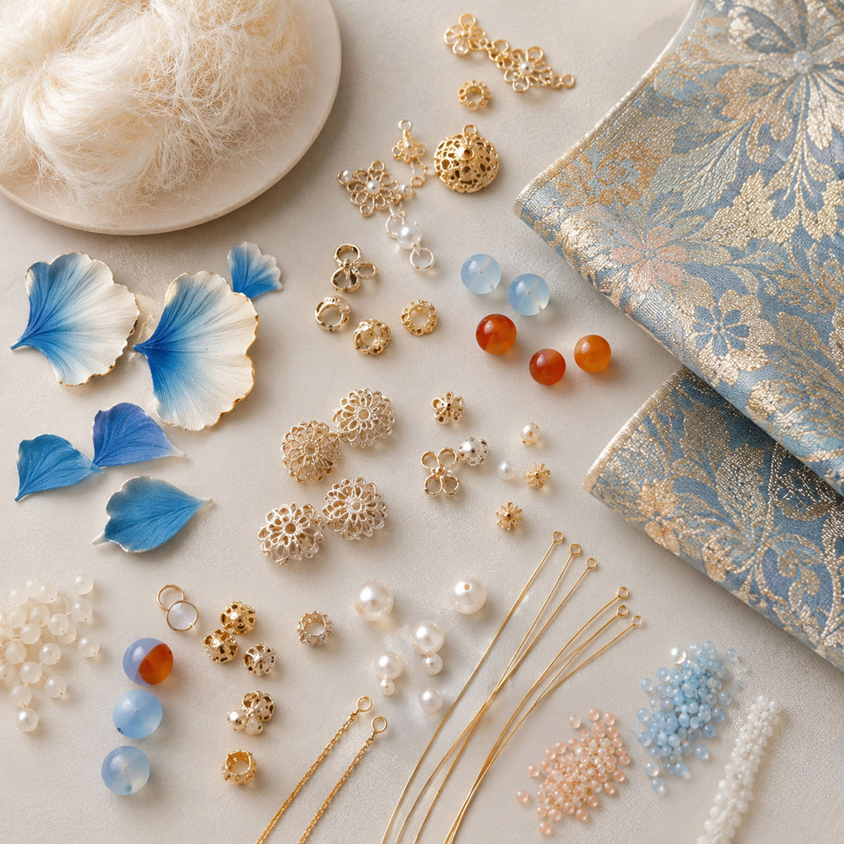 Fibres, blue floral forms, patterned fabric, beads and metal findings arranged on a light surface.